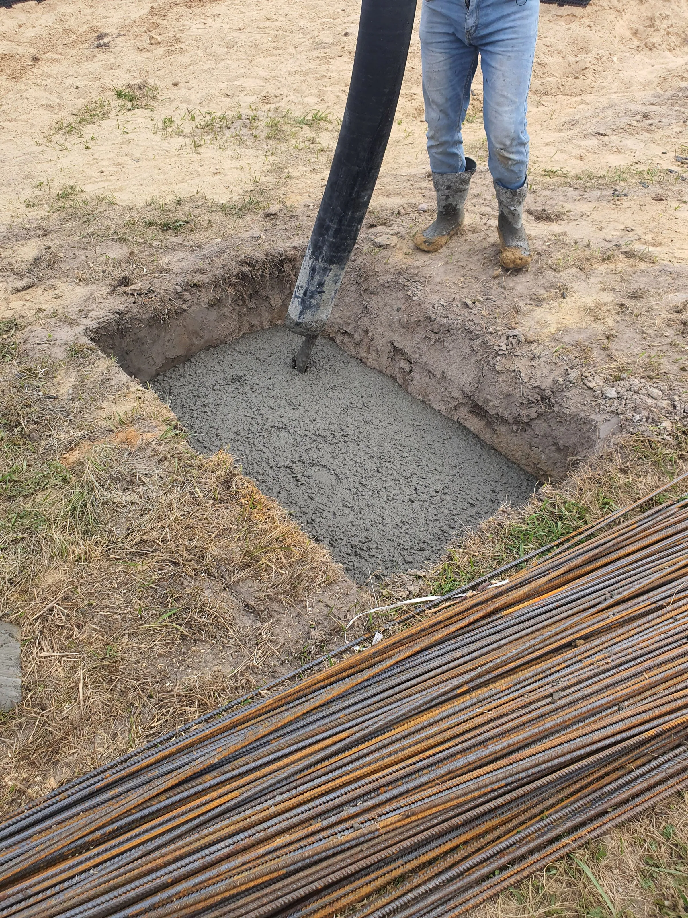 Wet concrete being pumped through a black hose into an excavated foundation pit, with bundled steel reinforcing bars on the ground nearby and a worker's boots at the edge