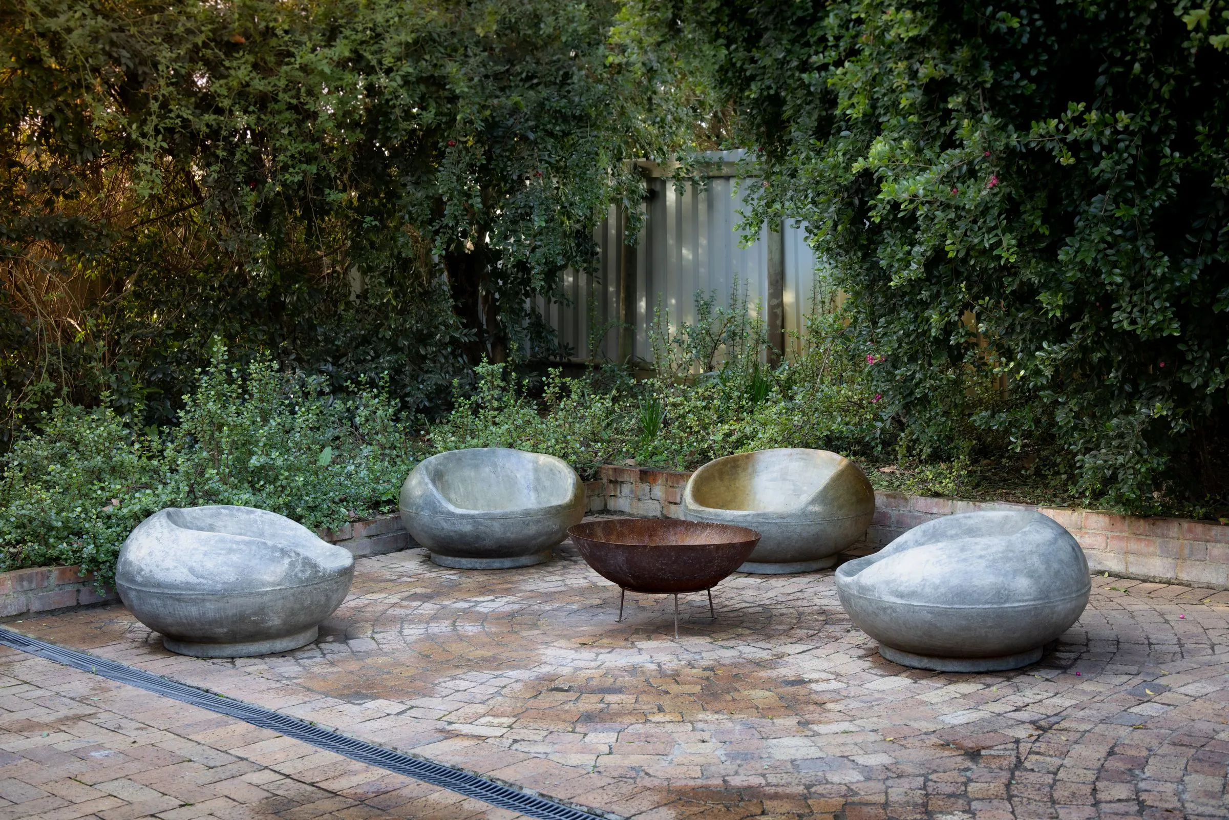 Outdoor patio with built stone bench seating arranged around a fire pit, surrounded by shrubs and trees