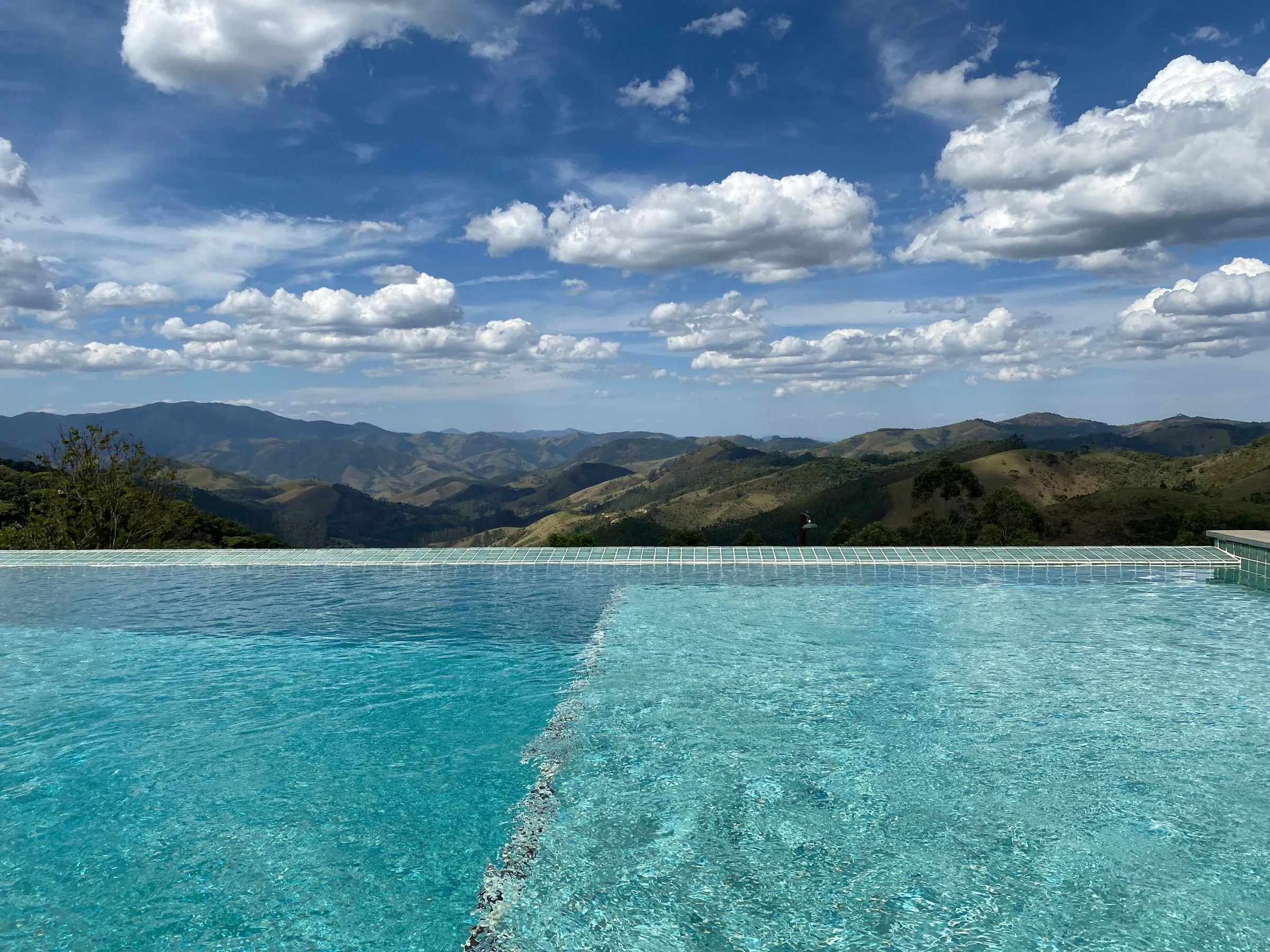 Water in a tiled pool meeting a level edge, with forested mountain ridges and a partly cloudy sky beyond