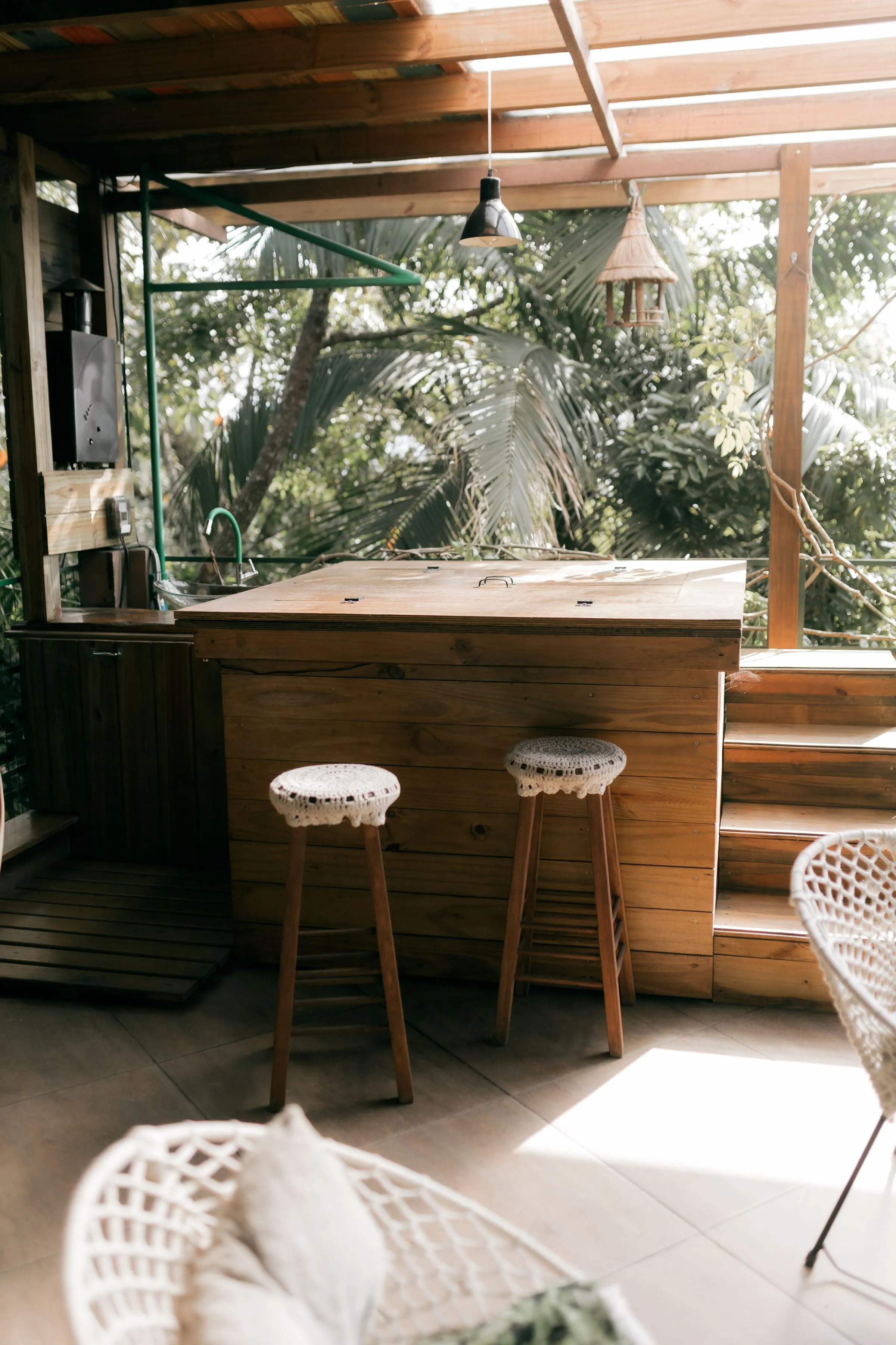 Timber bar counter with two woven-topped stools in an open-sided covered space, with a sink and pendant light and palm foliage outside