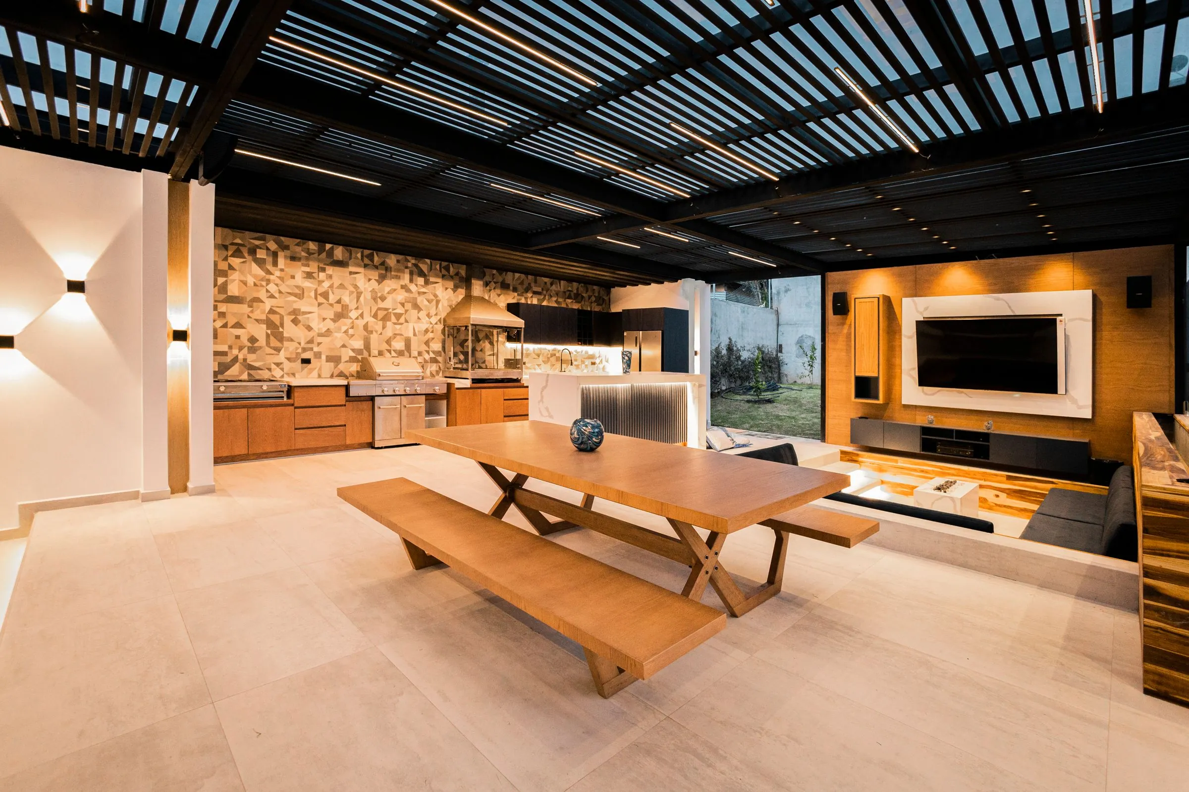 Wide covered outdoor kitchen and living space with a slatted timber ceiling, long wooden table with benches, run of cabinetry and a wall-mounted television