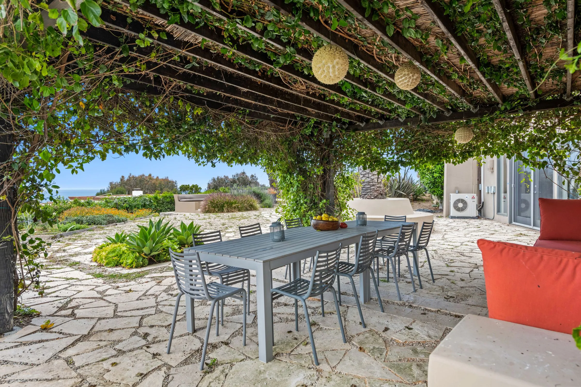 Outdoor dining table and chairs on a garden patio beneath a vine-covered pergola, with a view towards the sea