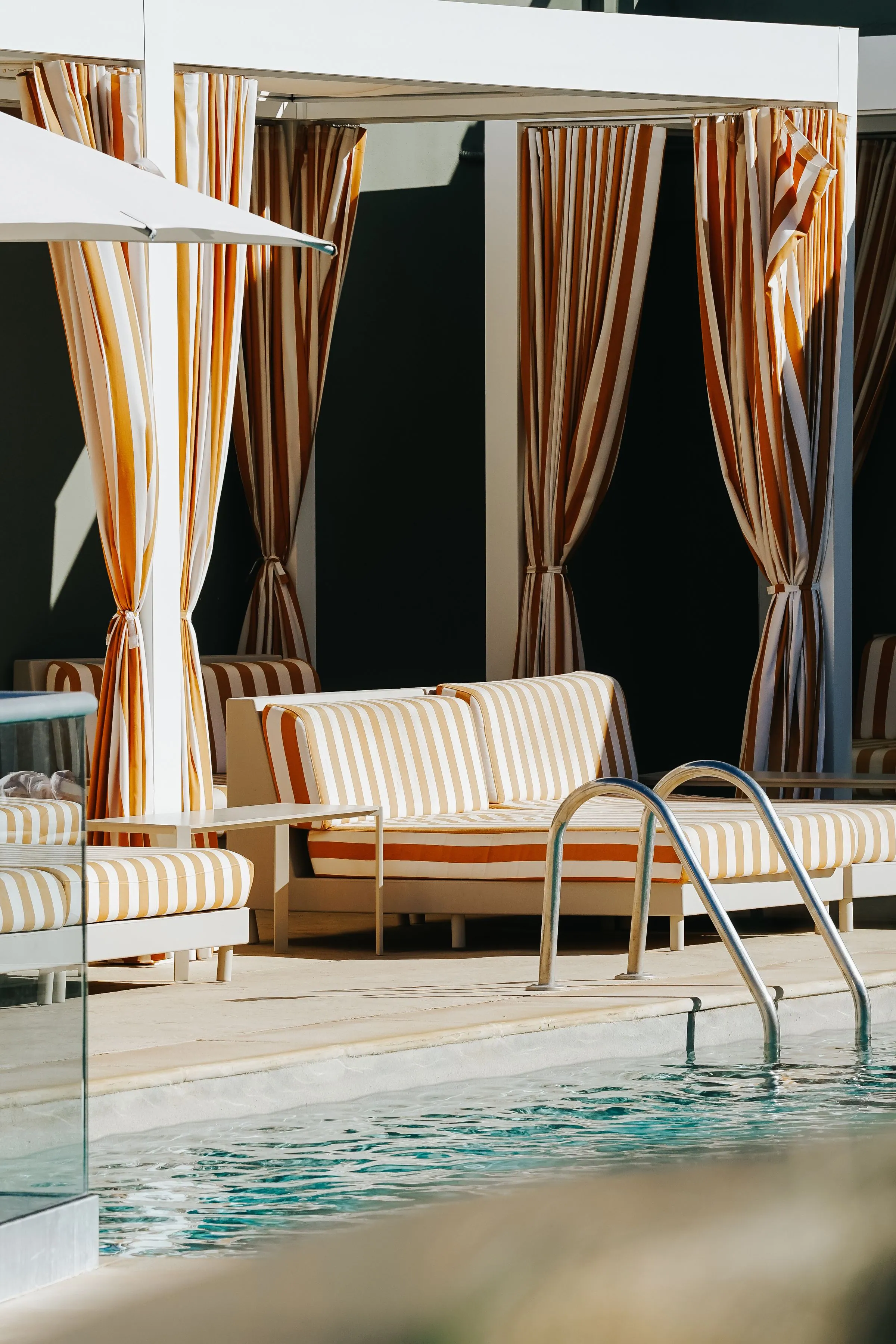 Poolside cabana with a fabric roof and striped side curtains, sun loungers set out beside the pool