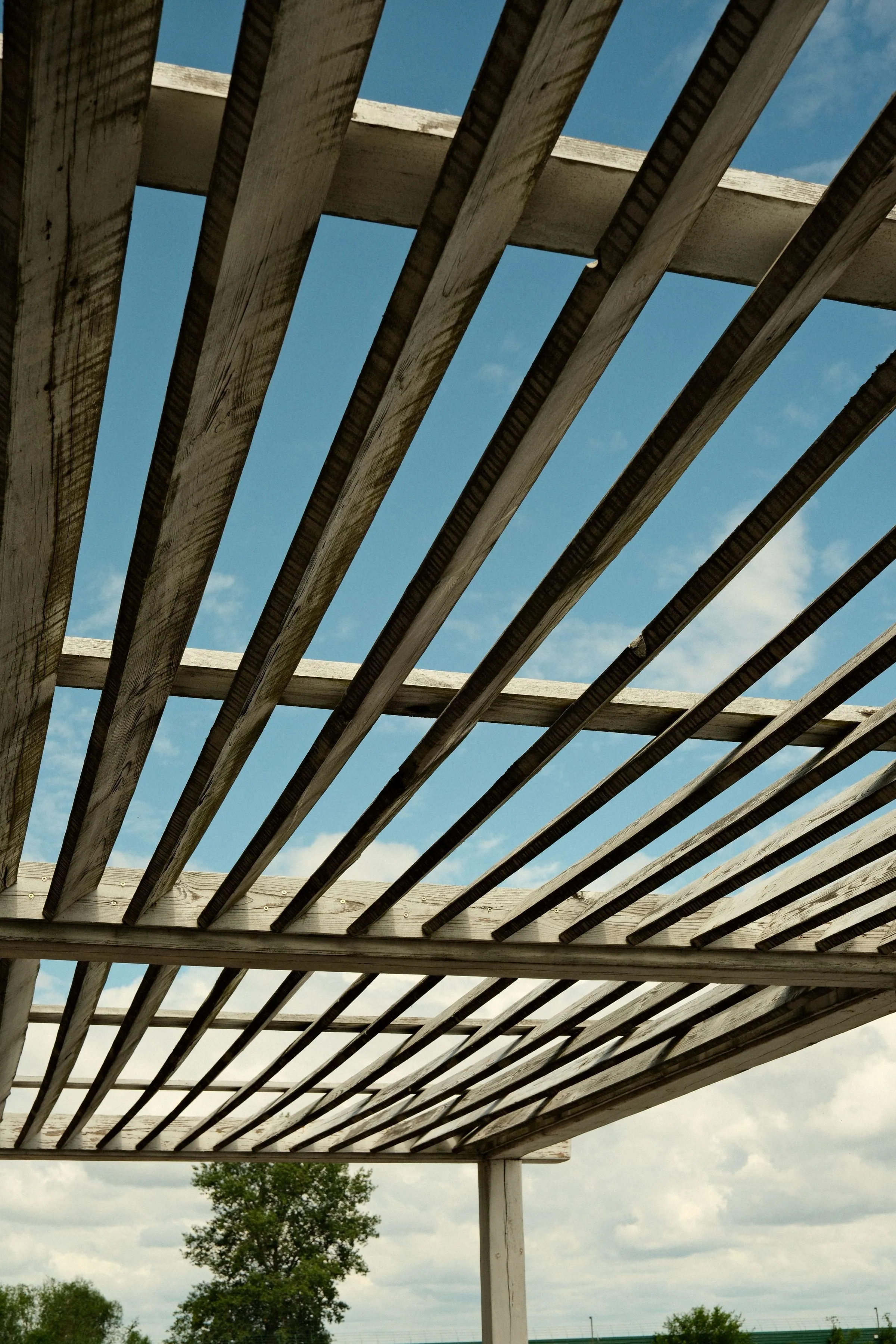 Looking up at a weathered timber pergola, its parallel slats and cross beams silhouetted against a blue sky with clouds