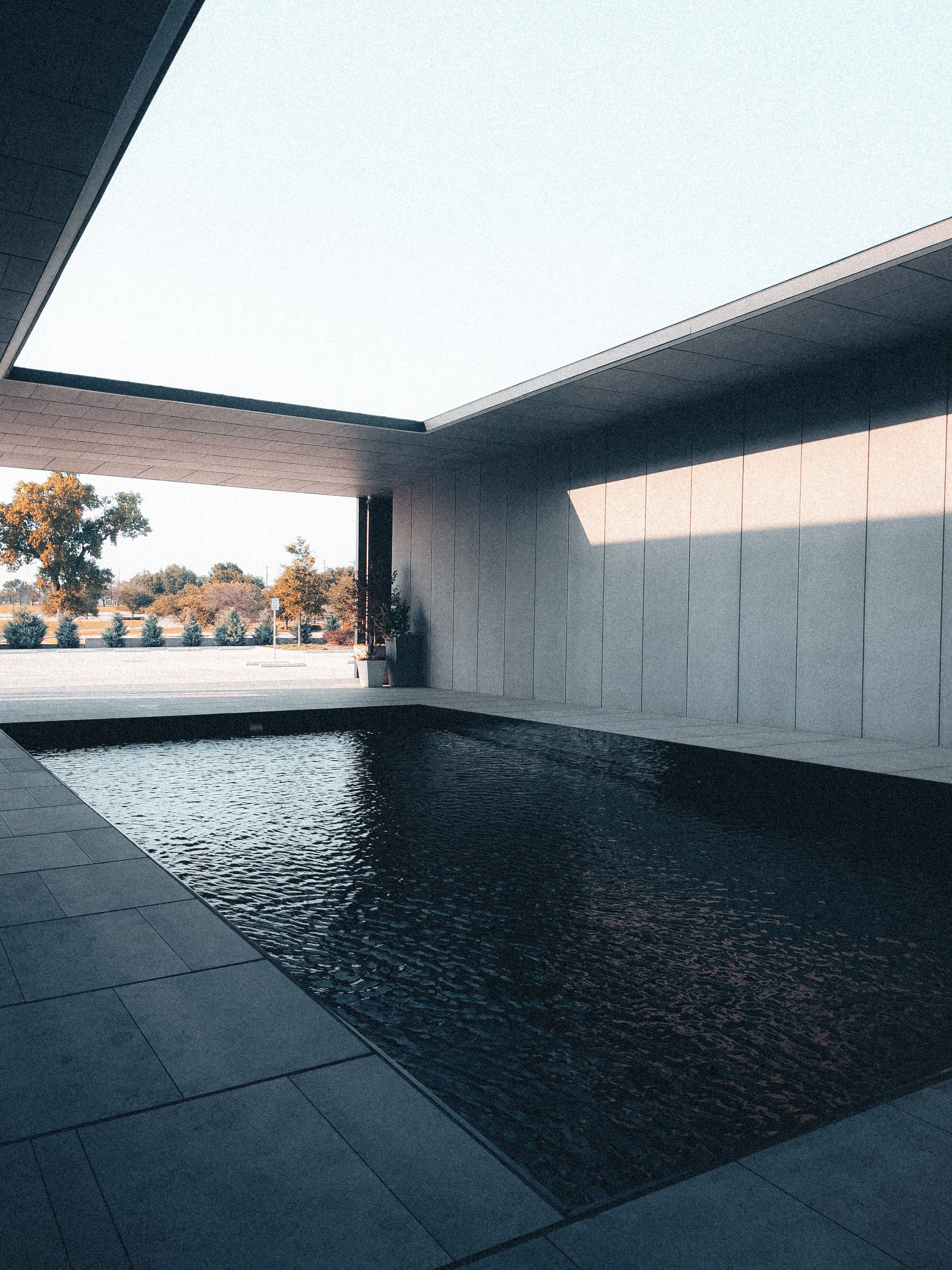 Rectangular pool with near-black rippled water and grey stone coping, set against a concrete-panelled wall and flat overhanging roof