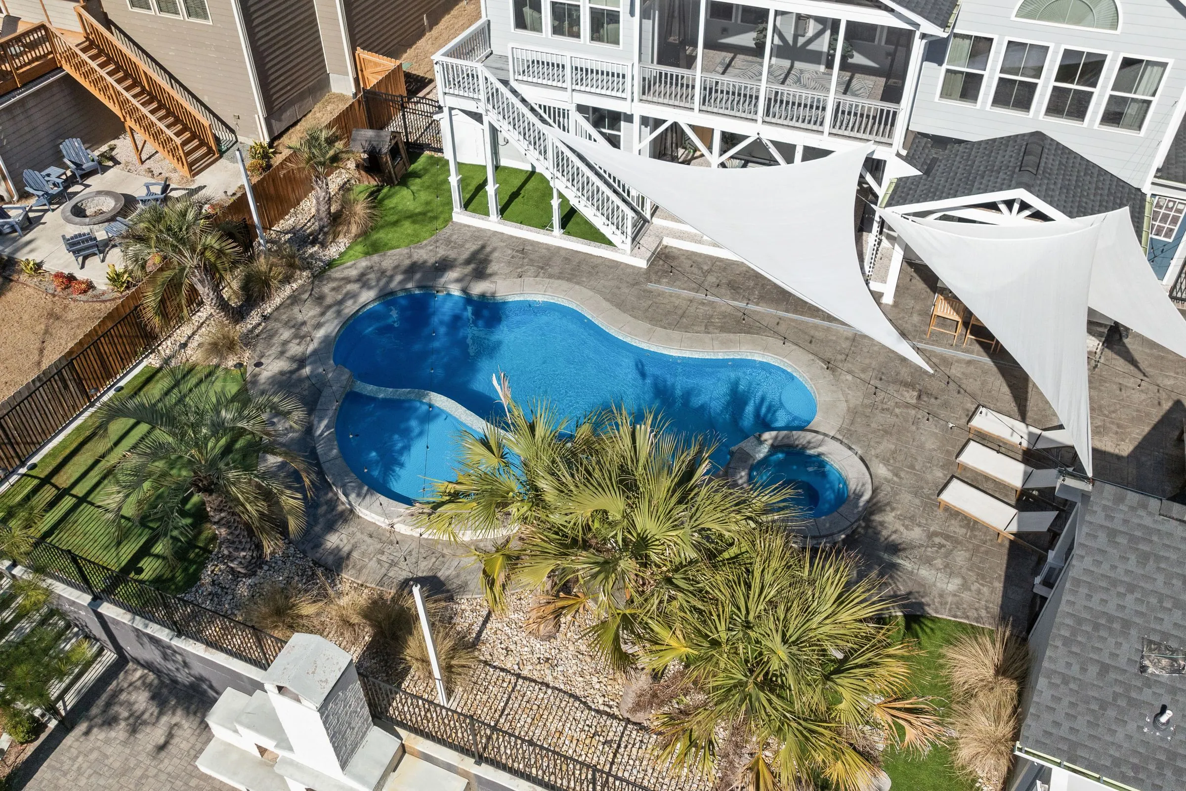 Aerial view of a curved freeform pool with an attached round spa, surrounded by paver decking, palms and shade sails behind a house