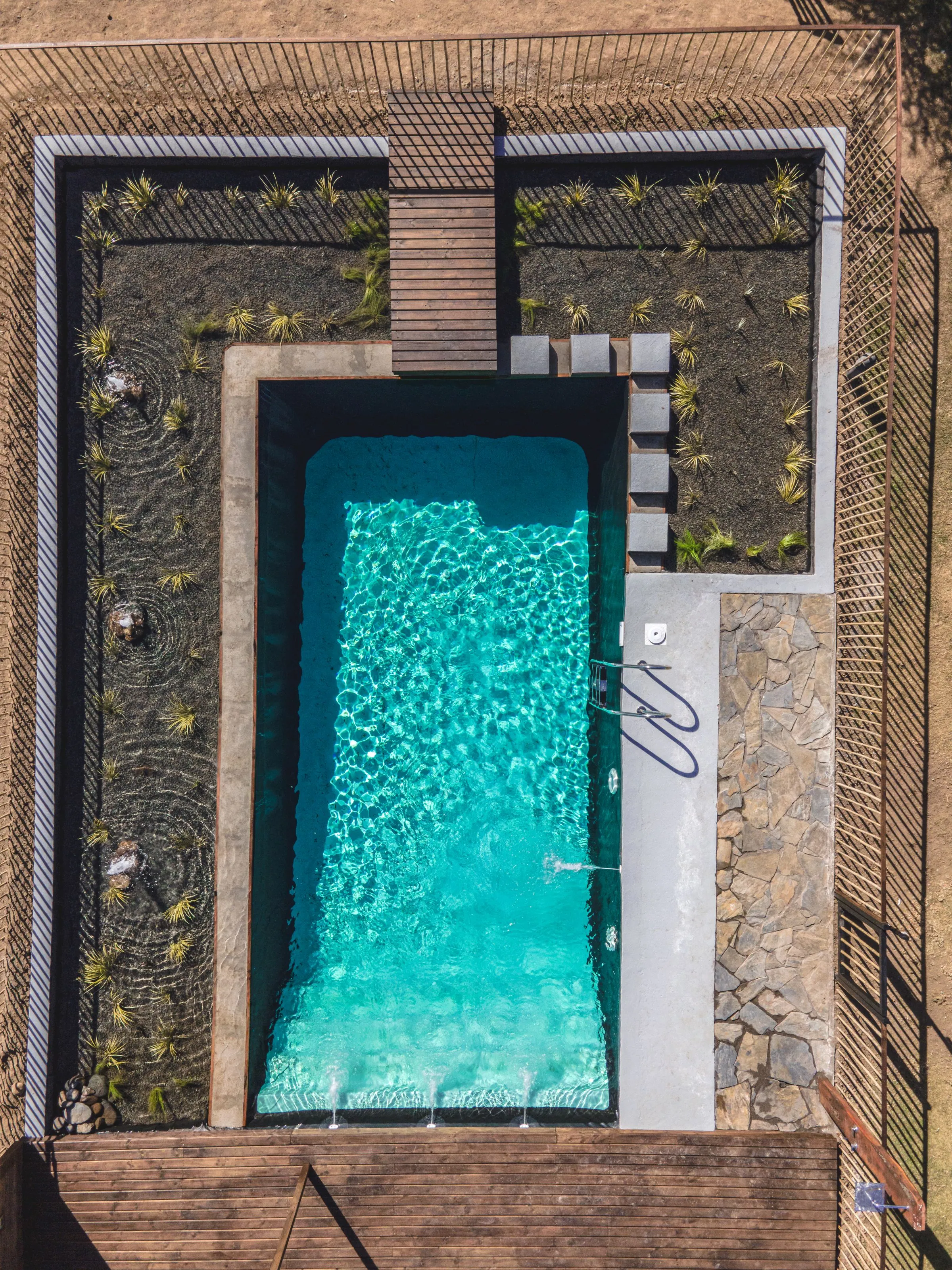 Overhead view of a rectangular pool set in timber decking, with square stepping stones, gravel planting beds and stone paving around it