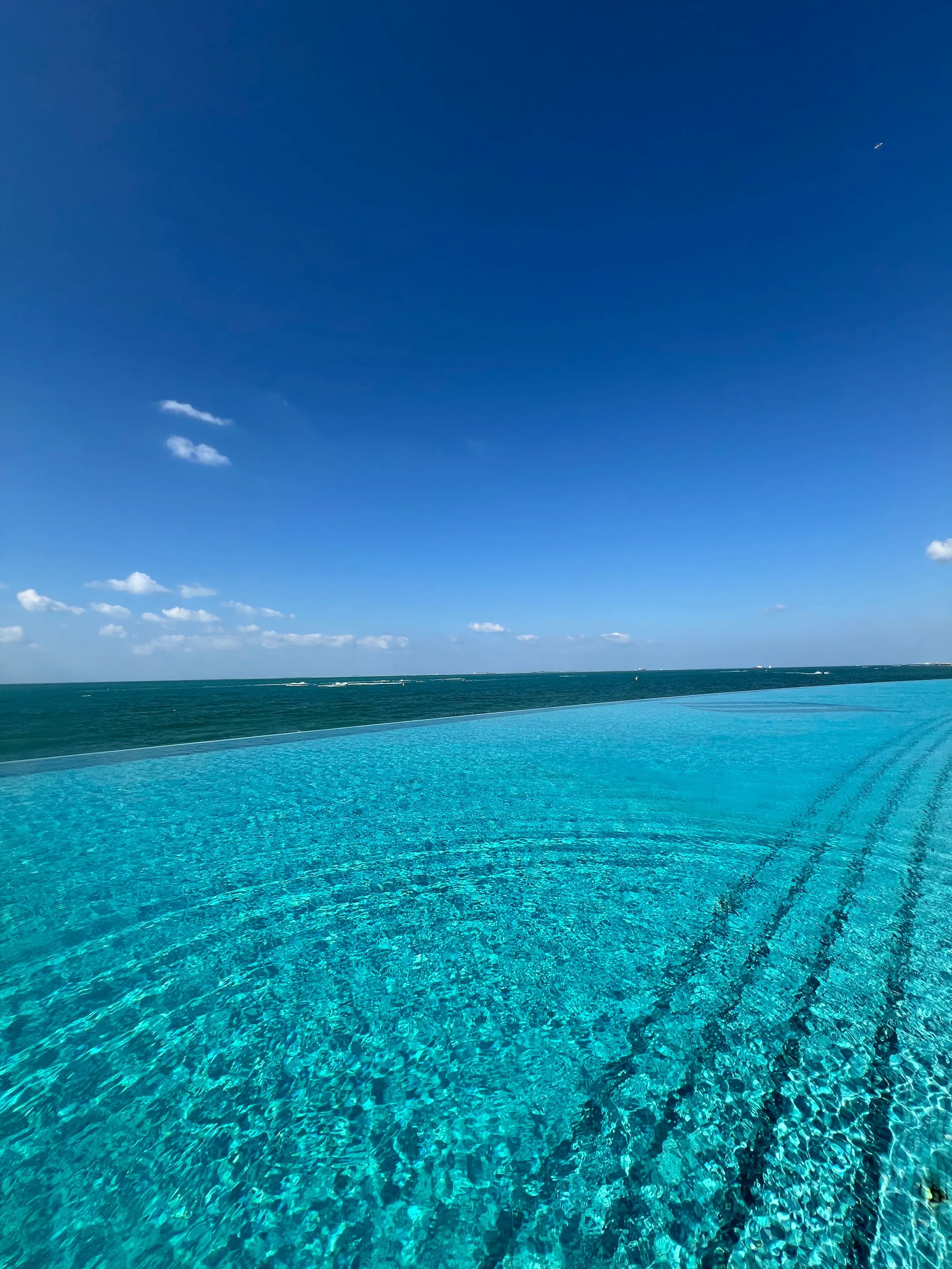 Turquoise pool water running to a level edge where it meets the darker sea, under a clear blue sky
