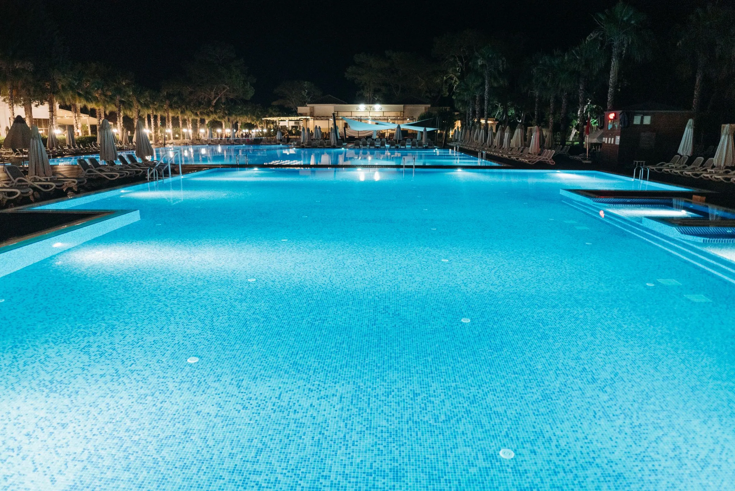 Large mosaic-tiled pool at night with underwater lights on, palm trees and lit buildings in the background