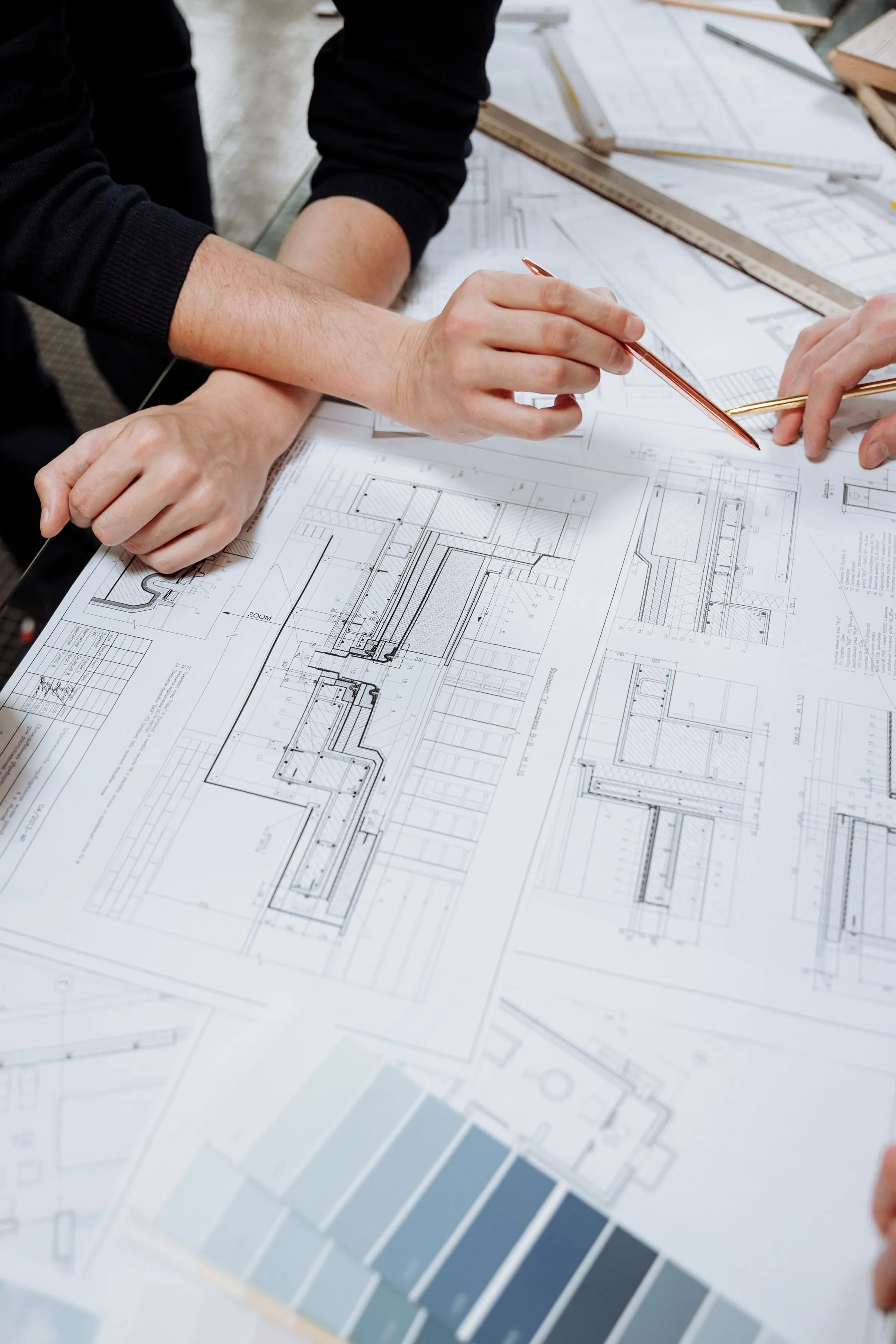 Two people leaning over large technical construction drawings on a table, one pointing with a pencil, with a metal ruler and a colour swatch fan alongside